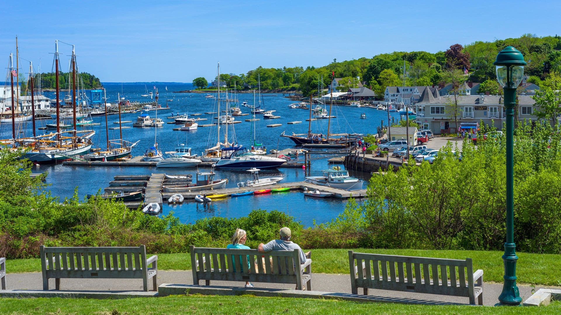 A couple sits on a bench overlooking a scenic harbor filled with boats and greenery.