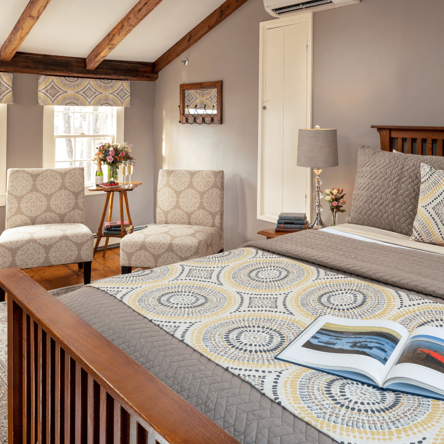 A cozy bedroom featuring a wooden bed, patterned bedding, two chairs, a small table with flowers, and a soft color palette.