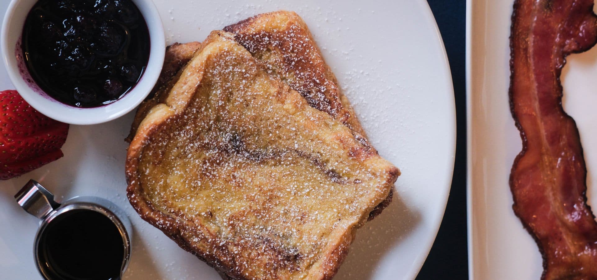 A plate featuring golden French toast dusted with powdered sugar, served with a side of berry compote and crispy bacon.