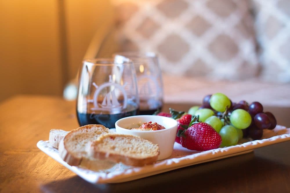 A platter with sliced bread, fruit, and a small dish, accompanied by two glasses of wine.