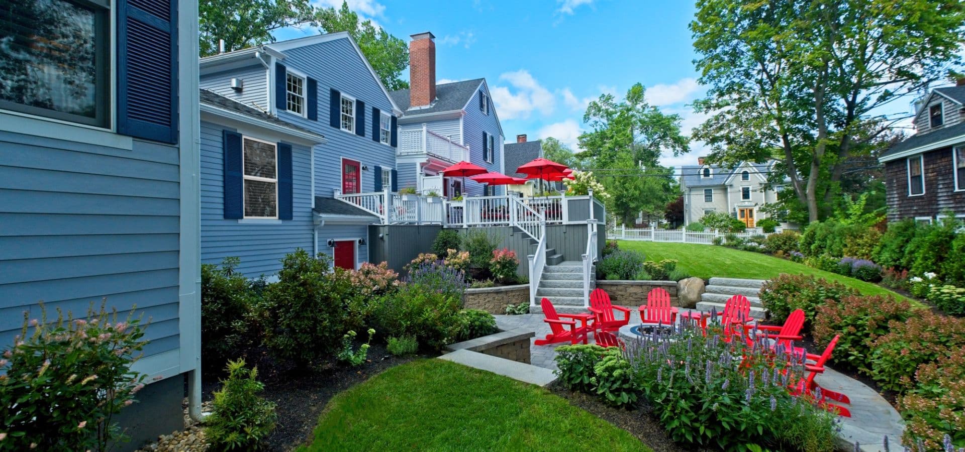 A well-maintained backyard features a patio with red umbrellas and chairs, surrounded by lush greenery and blue houses.