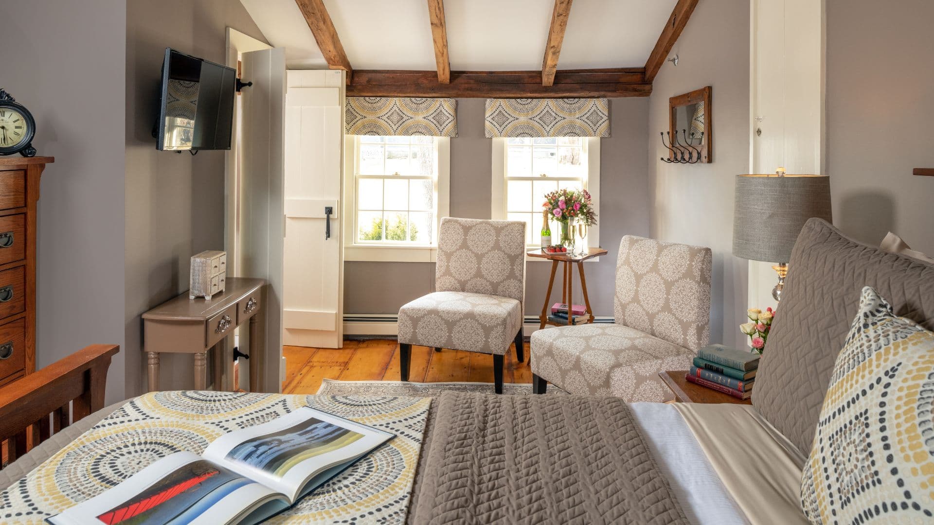 A cozy bedroom featuring a bed with decorative pillows, two armchairs, a small table, and large windows with flower arrangements.