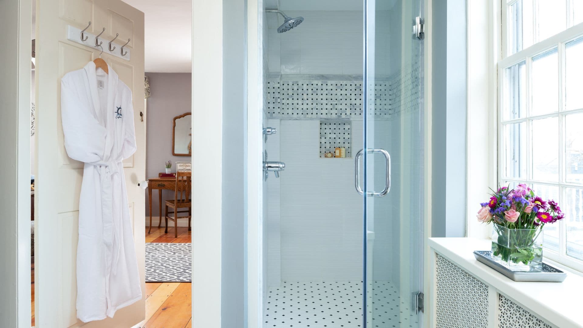 Bright bathroom featuring a glass shower, a hanging white robe, and a bouquet of flowers by the window.