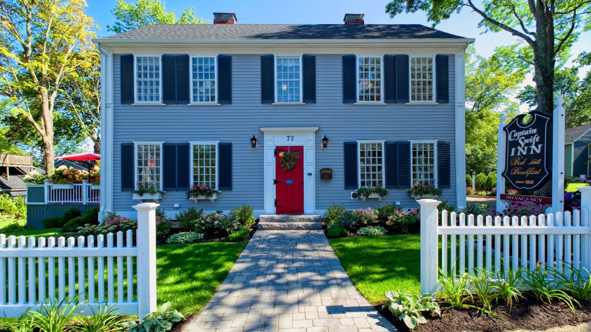 A charming blue inn with a red door surrounded by greenery and a white picket fence.